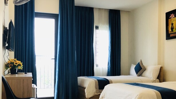 Ein Hotelzimmer mit zwei Betten, einem Schreibtisch mit Stuhl, einem Standventilator und einem großen Fenster mit Vorhängen.