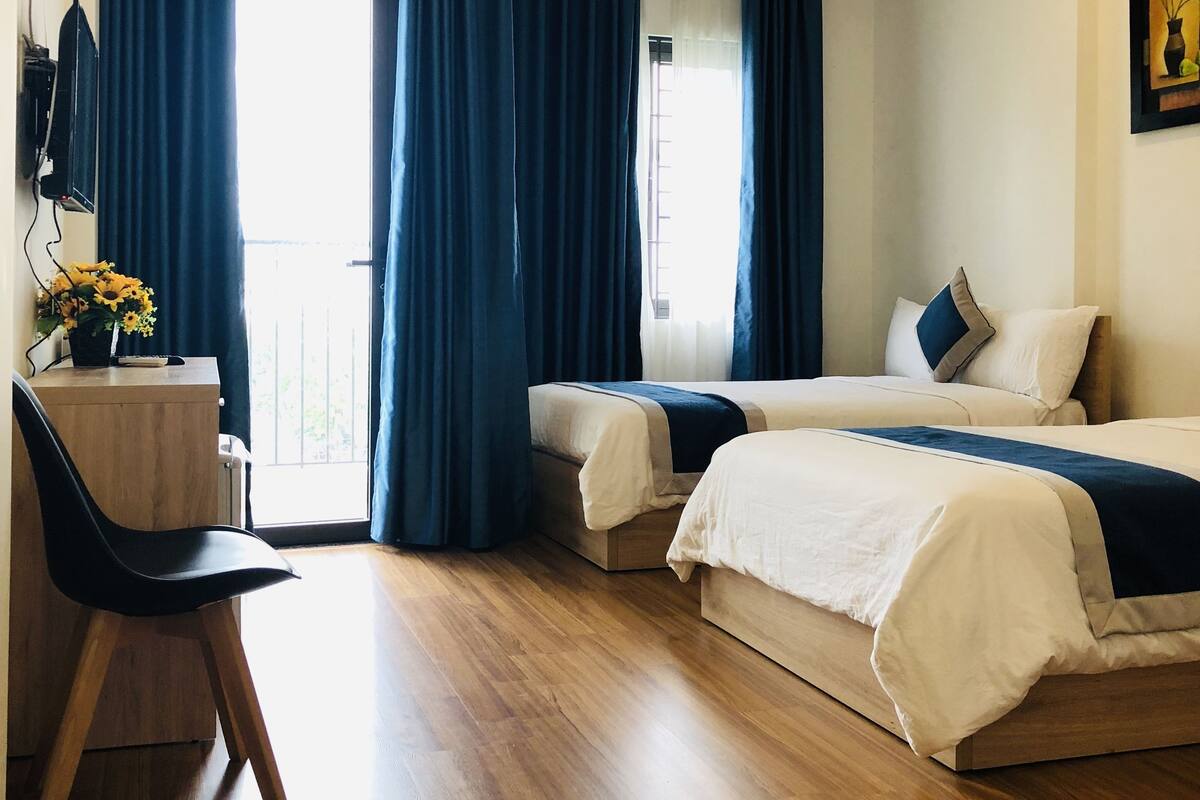 A hotel room with two beds, a desk with a chair, a standing fan, and a large window with curtains.