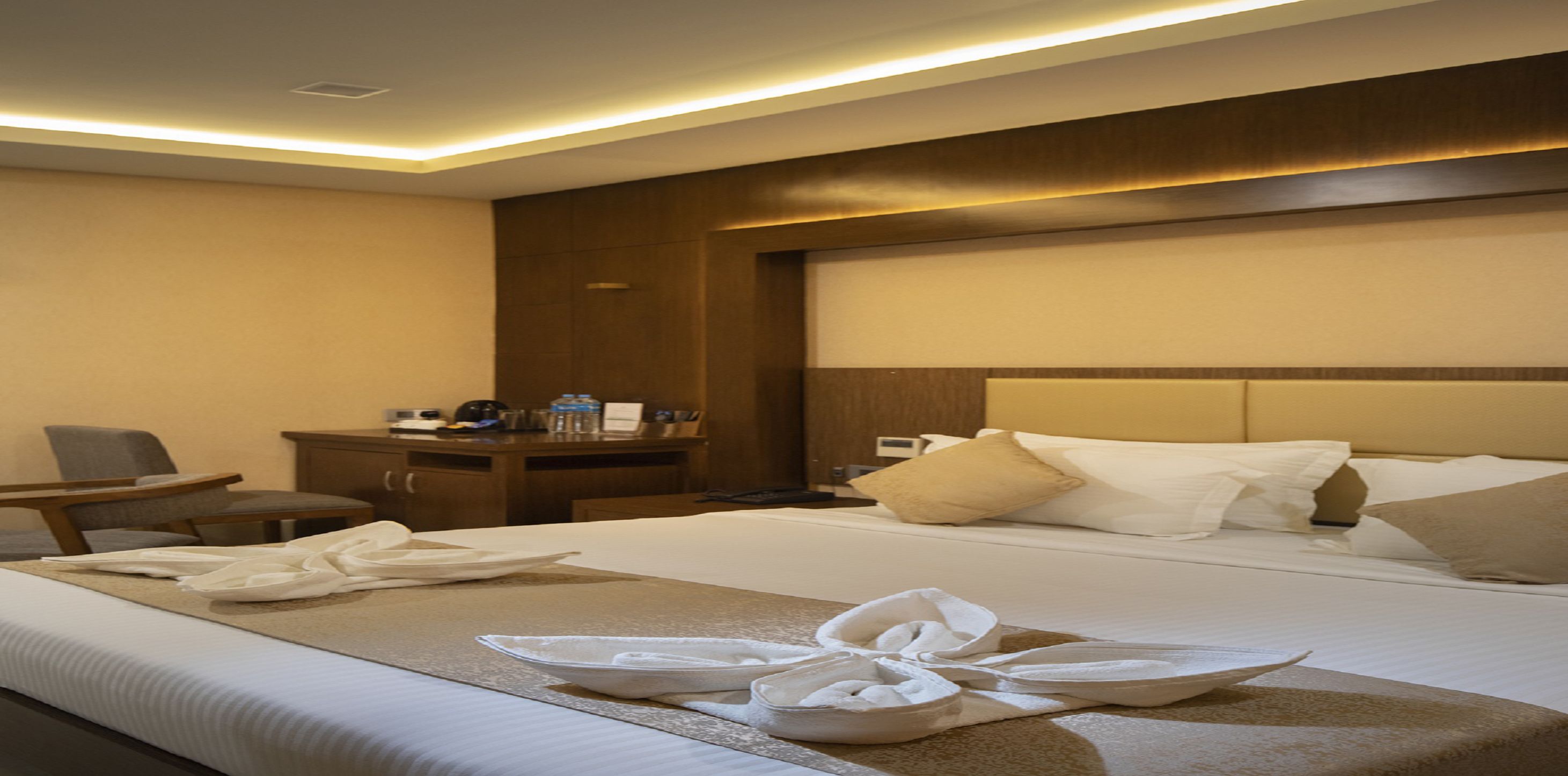 A hotel room with a neatly made bed, a desk with a chair, and a bedside table with a lamp and a bottle of water.