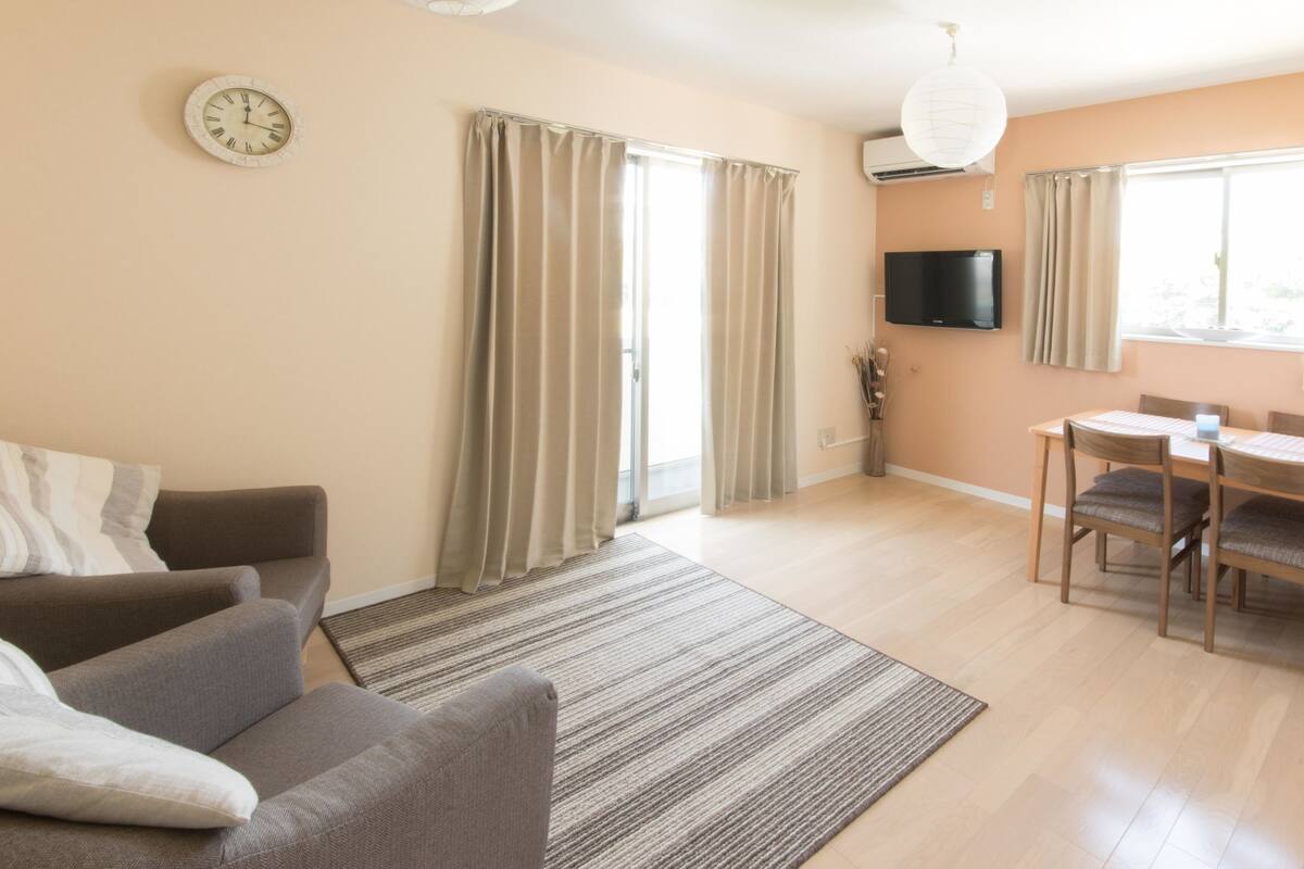 A living room with a sofa, a dining table with chairs, a TV mounted on the wall, and a large window with curtains.