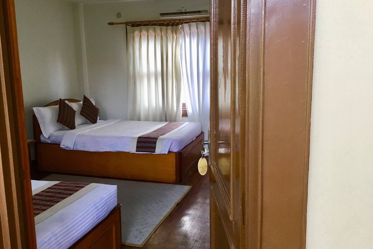 A bedroom with two beds, a wooden headboard, a bedside table, a nightstand, a window with curtains, and a ceiling fan.