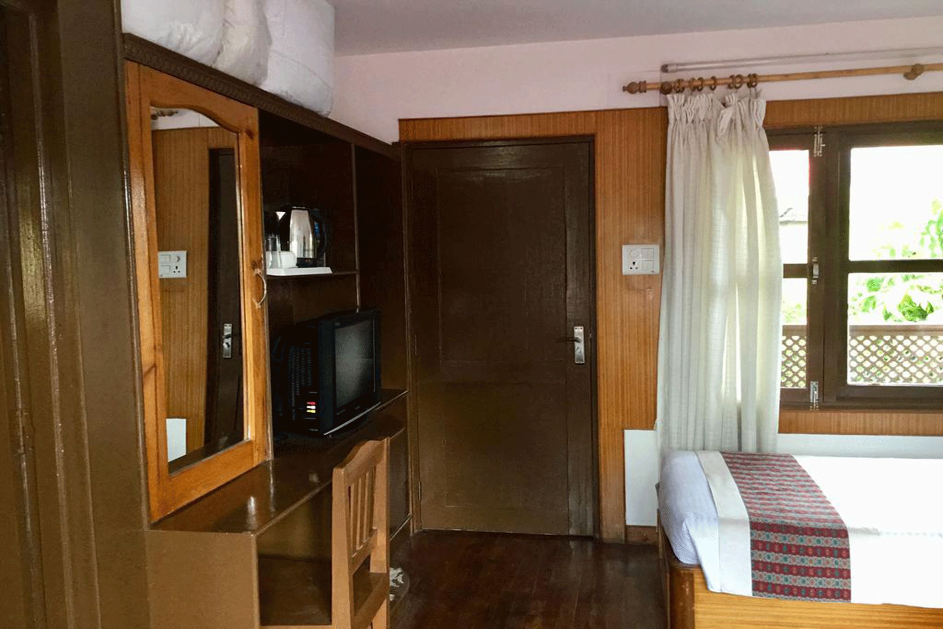 A room with a bed, a television on a wooden shelf, a door, and a window with a curtain.