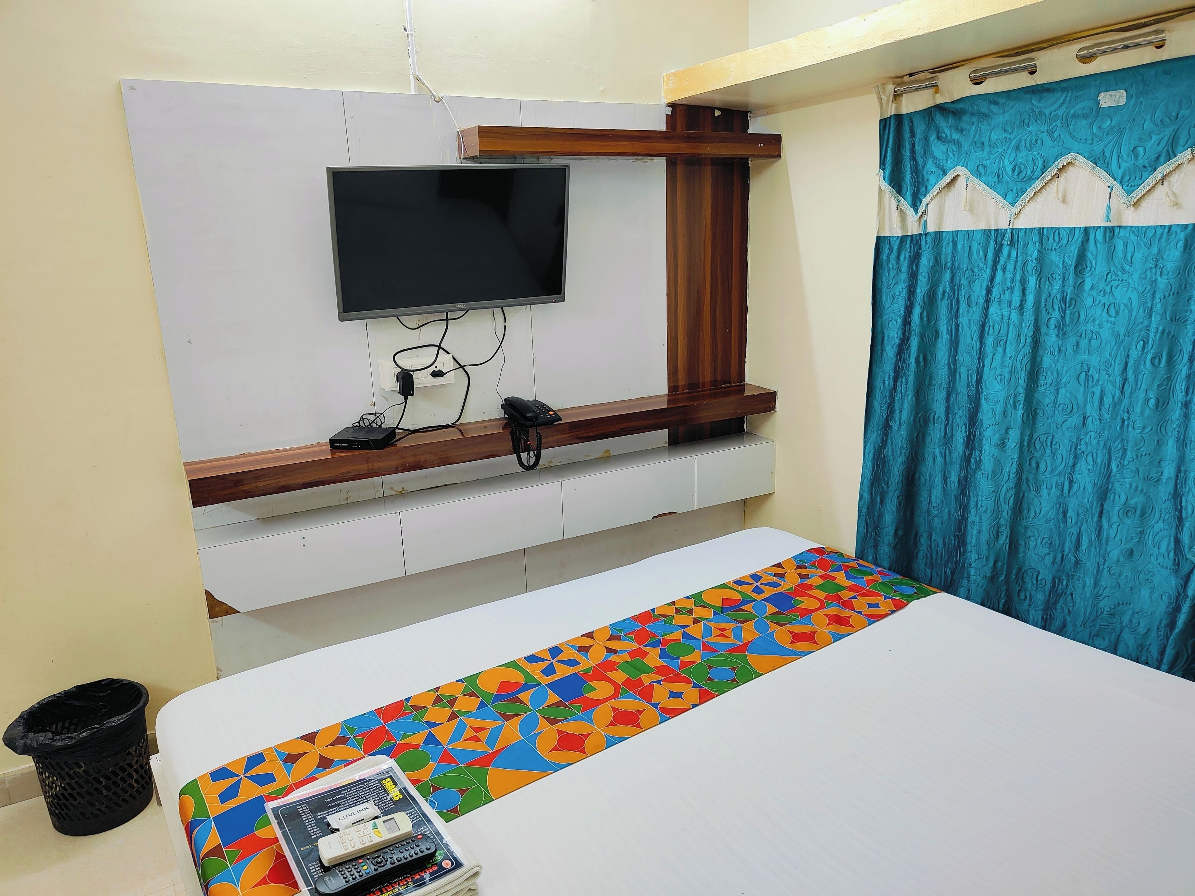 A hotel room with a bed, a TV mounted on the wall, a wooden shelf, a curtain, and a trash bin.