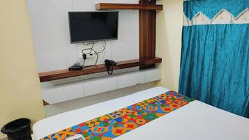 A hotel room with a bed, a TV mounted on the wall, a wooden shelf, a curtain, and a trash bin.