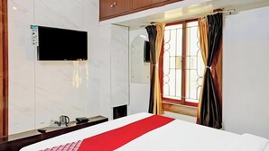 Ein Hotelzimmer mit einem Bett, einem an der Wand montierten Fernseher, einem Fenster mit Vorhängen und einem Nachttisch über dem Bett.