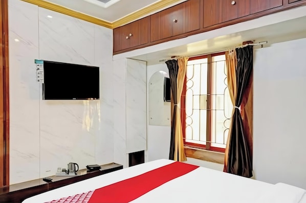 Habitación de hotel con cama, televisor montado en la pared, ventana con cortinas y un armario de madera sobre la cama.