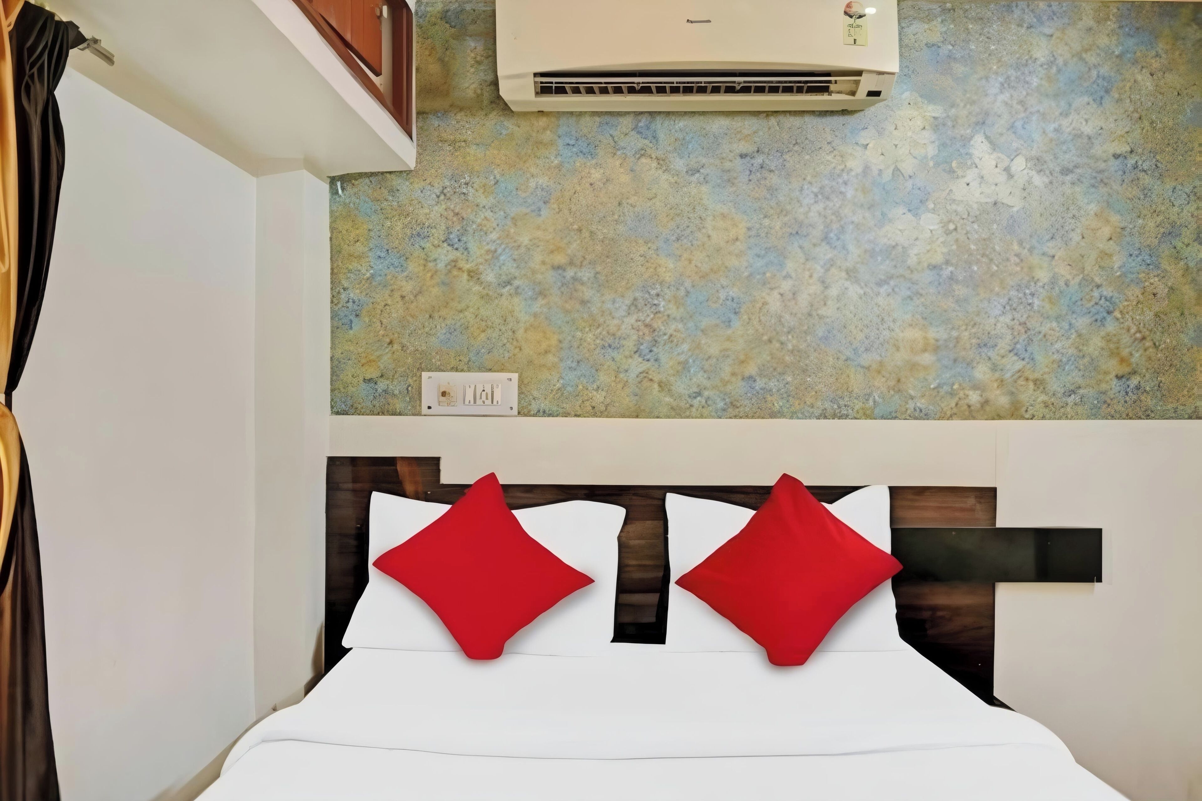 Habitación de hotel con una cama, dos almohadas rojas, aire acondicionado montado en la pared y un panel de pared con textura.