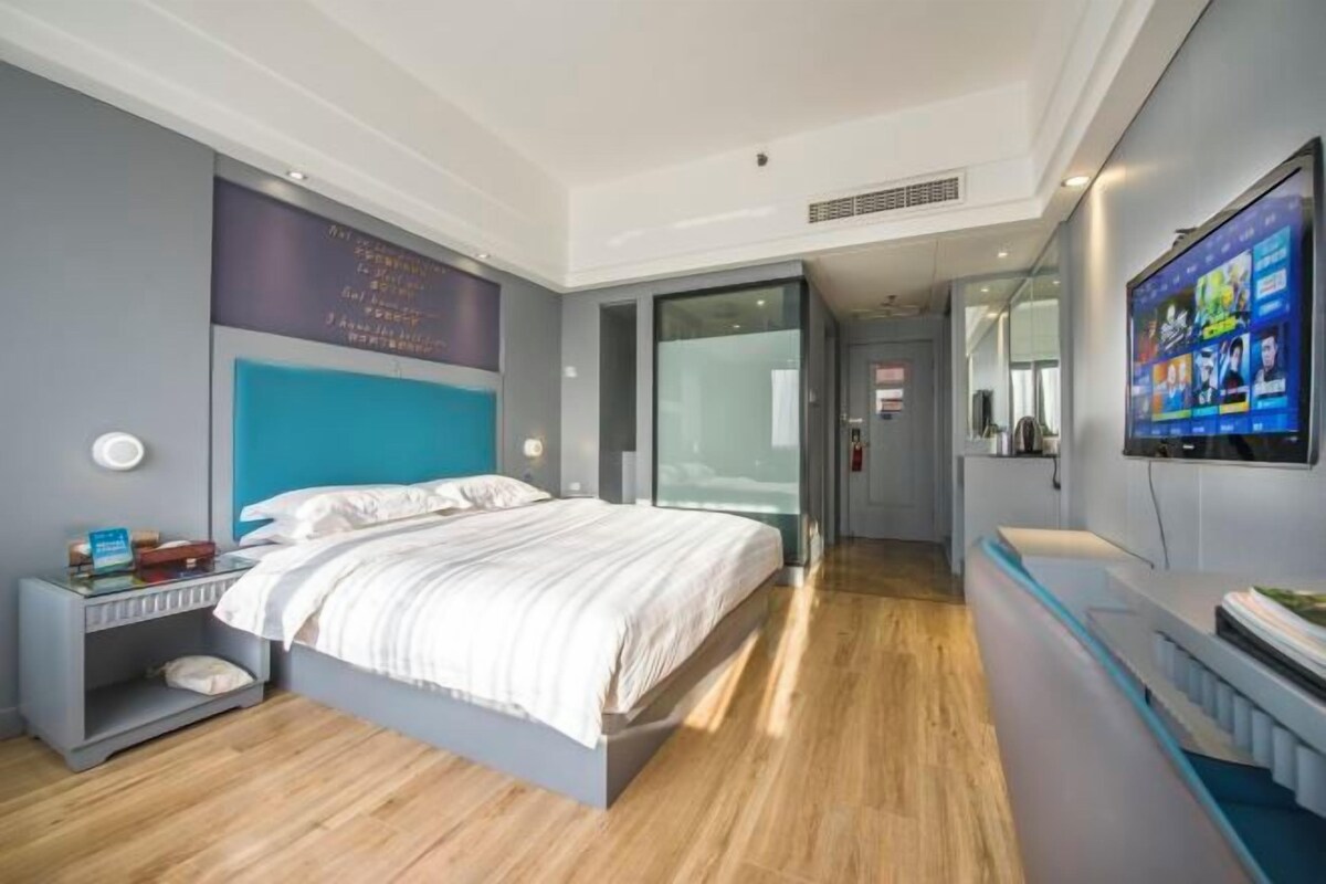 A modern hotel room with a bed, a TV, a desk, and a glass partition.