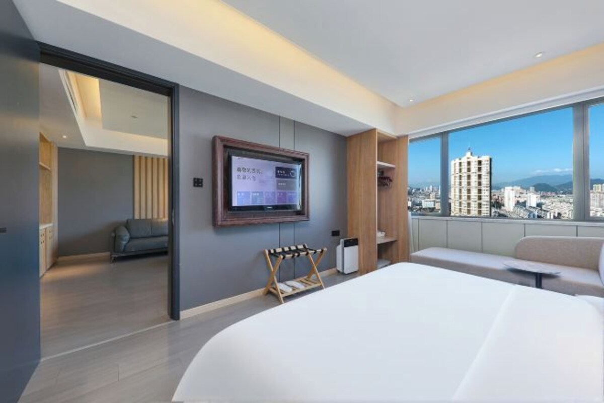 Una habitación de hotel moderna con una cama grande, un televisor montado en la pared, una zona de estar con vista a la ciudad y un balcón con vistas panorámicas.