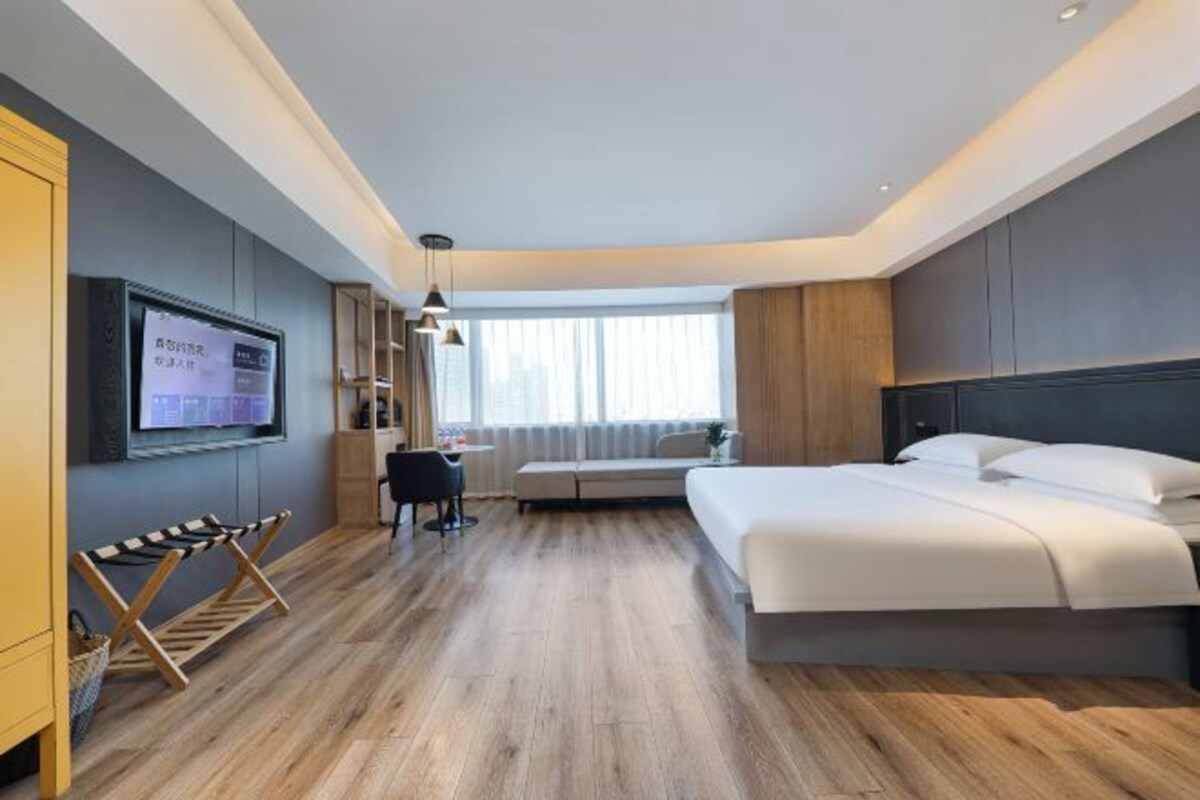 Una habitación de hotel moderna con una cama grande, un escritorio con silla, un televisor montado en la pared y un ventanal con cortinas.