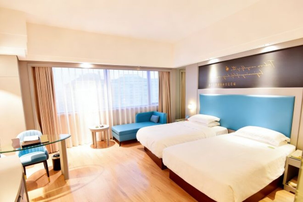 Habitación de hotel con dos camas, un pequeño rincón de estar, una mesa de vidrio y vistas a la ciudad a través de amplias ventanas.