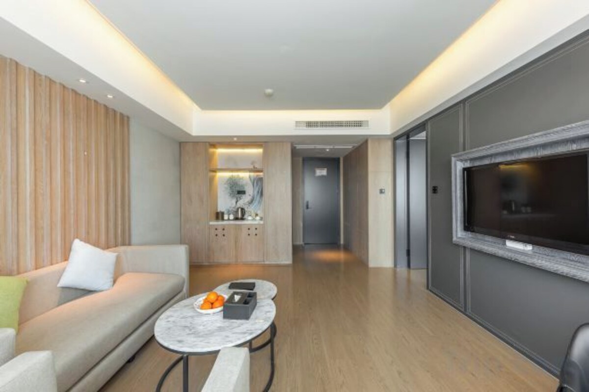 A modern hotel room with a sofa, a round coffee table, a flat-screen TV, and a kitchen area in the background.