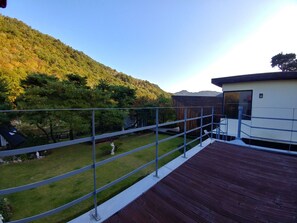 A wooden deck with a metal railing overlooking a grassy yard and a forested hillside. - Biuum Pension (Pyeongchang)