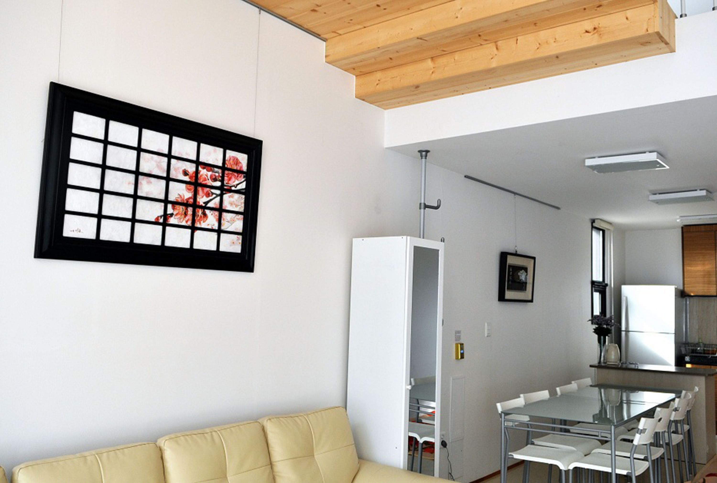 A modern living room with a beige sofa, a glass dining table with chairs, a kitchen area with a refrigerator, and a framed artwork on the wall.