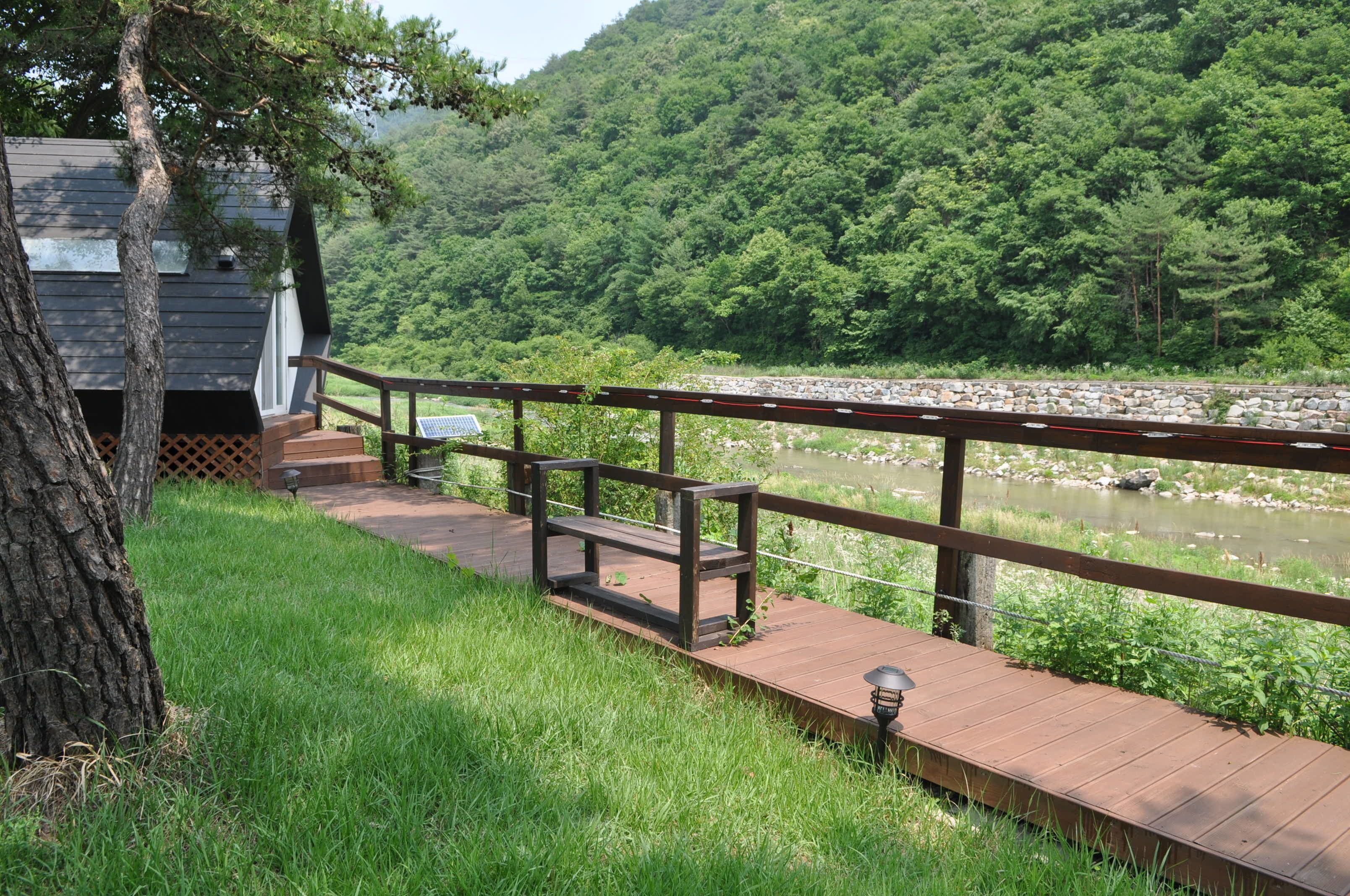 A wooden deck with a railing overlooking a river, surrounded by lush greenery and a hill in the background.