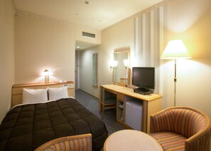 Quarto de hotel com uma cama, uma escrivaninha com televisão, uma cadeira e um abajur.