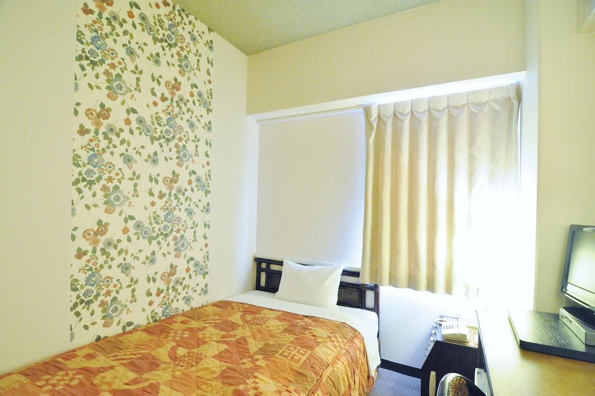 Une chambre d’hôtel avec un lit, un bureau avec un ordinateur, une fenêtre avec des rideaux et un papier peint à motifs floraux.