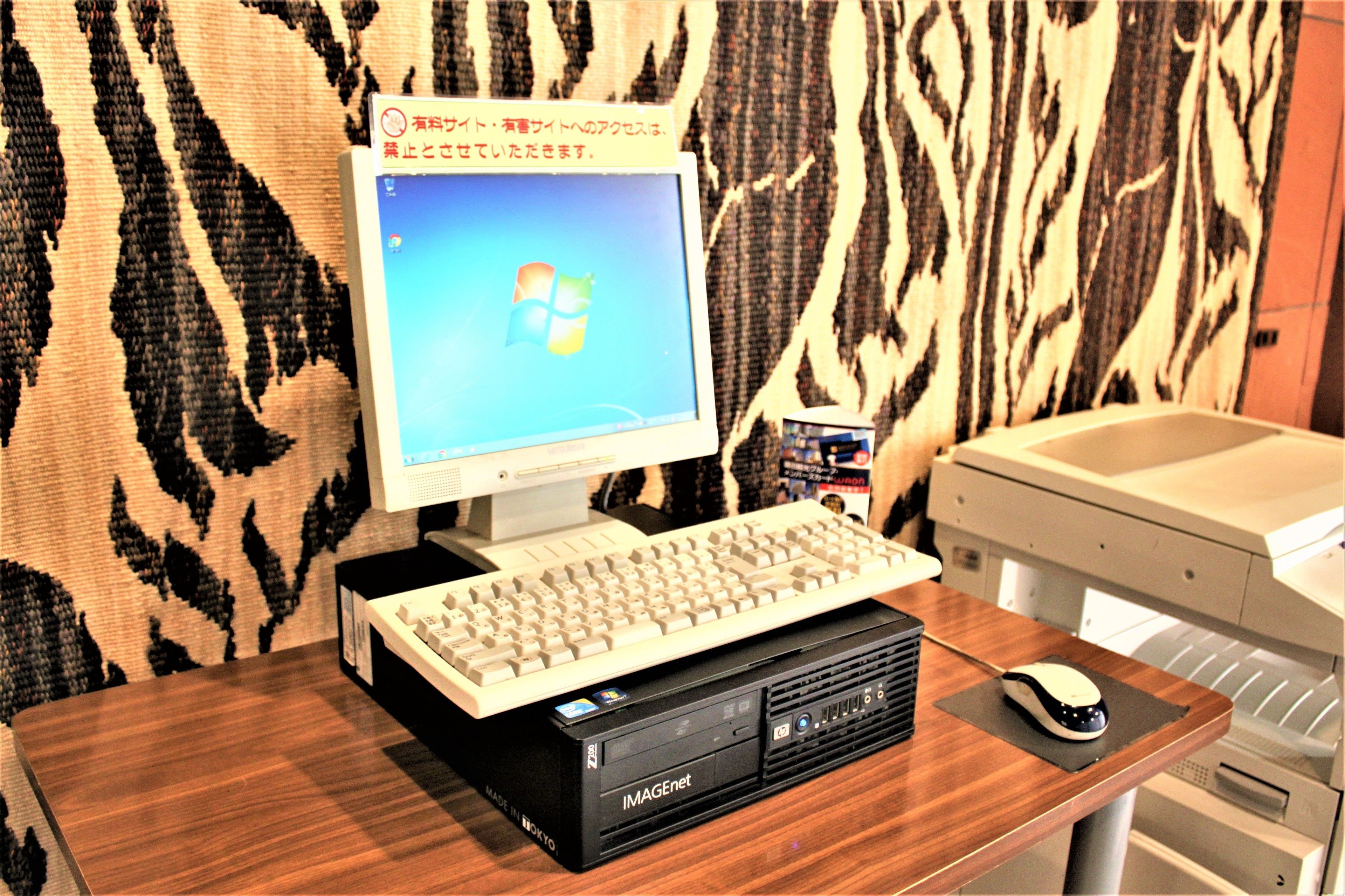 A desktop computer with a keyboard and mouse on a wooden desk, a printer, and a patterned wall in the background.