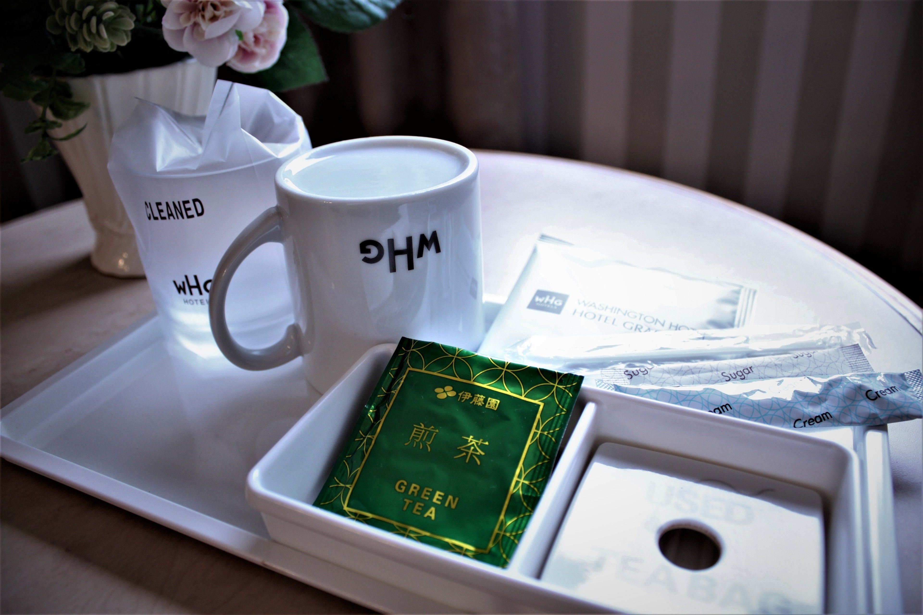 Una bandeja con una taza blanca, un sobre de té verde y una bolsa de tela con la etiqueta 'CLEANED' sobre una mesa blanca.