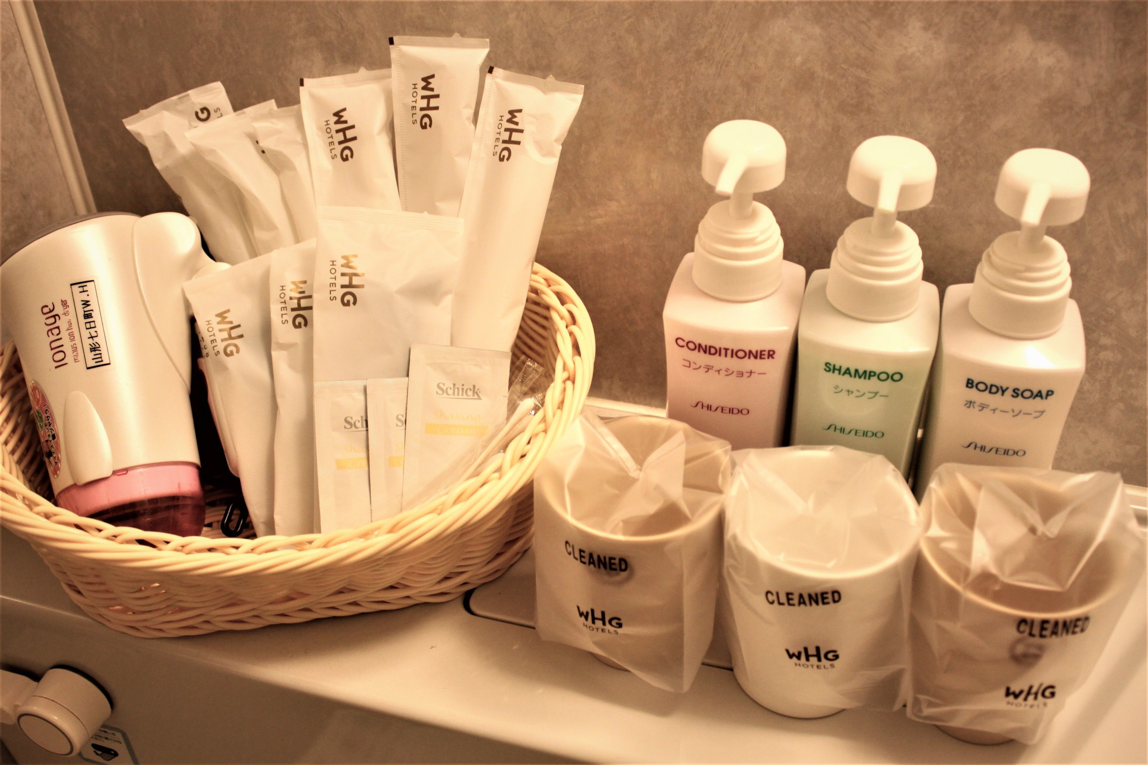 Een badkamer met toiletartikelen, waaronder een mandje met WHG hotelproducten, een fles conditioner en verschillende flessen shampoo en douchegel.