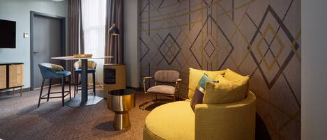 A modern hotel room with a round yellow sofa, a dining area with a table and chairs, a TV mounted on the wall, and a patterned wallpaper.
