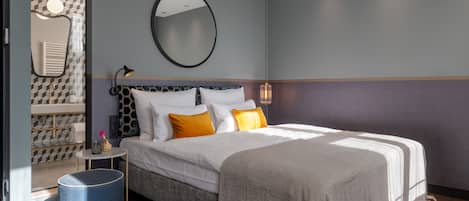 A modern hotel room with a large bed, a round mirror, a small side table, and a blue ottoman.