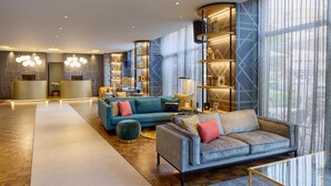 A spacious lobby with a central seating area featuring a grey sofa and a teal sofa, surrounded by wooden flooring and a large window with sheer curtains. - Postboutique Hotel (Wuppertal)