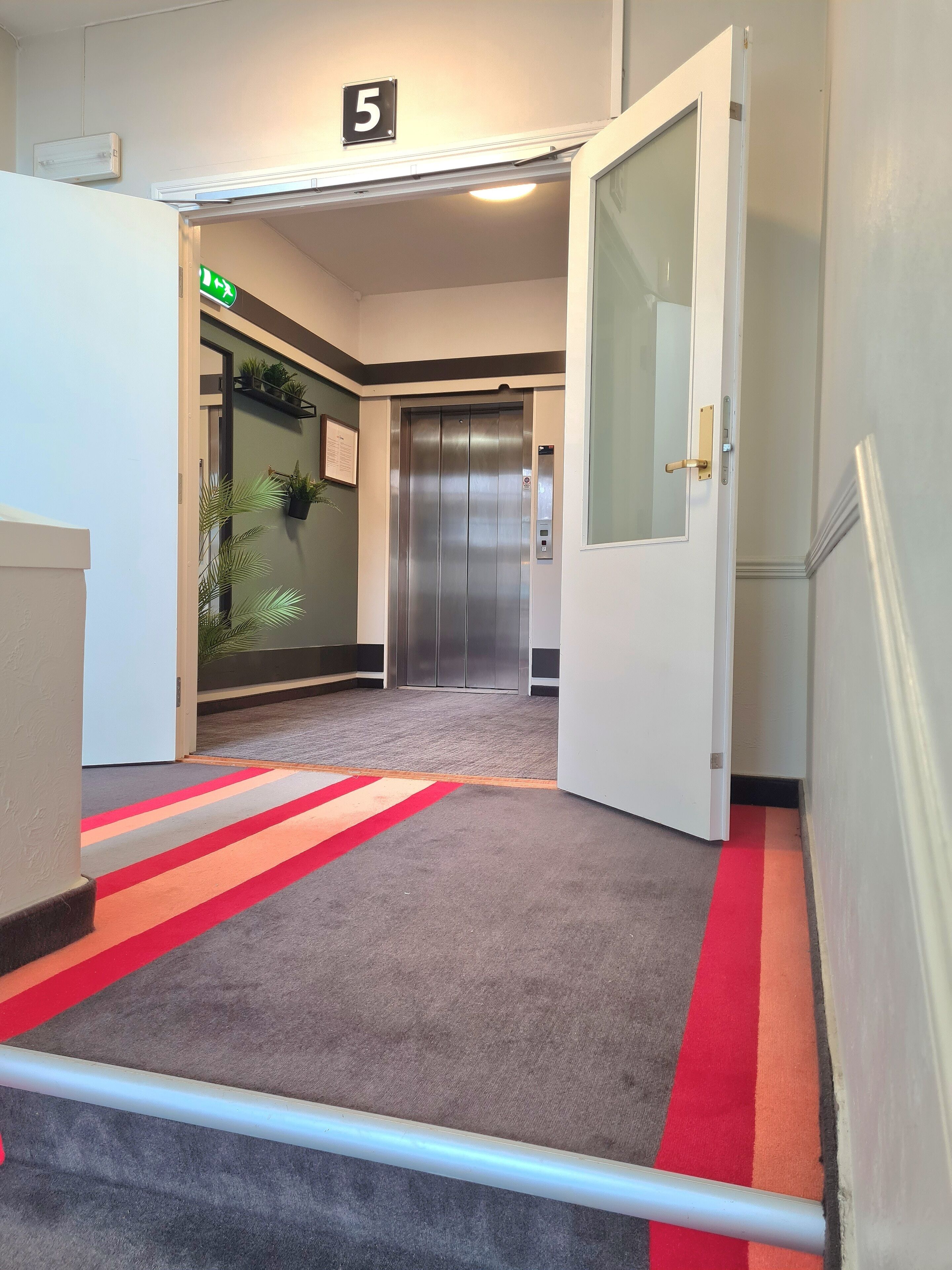 Un ascenseur aux finitions métalliques, une porte dotée d’une poignée et un sol recouvert de moquette à rayures rouges et grises.