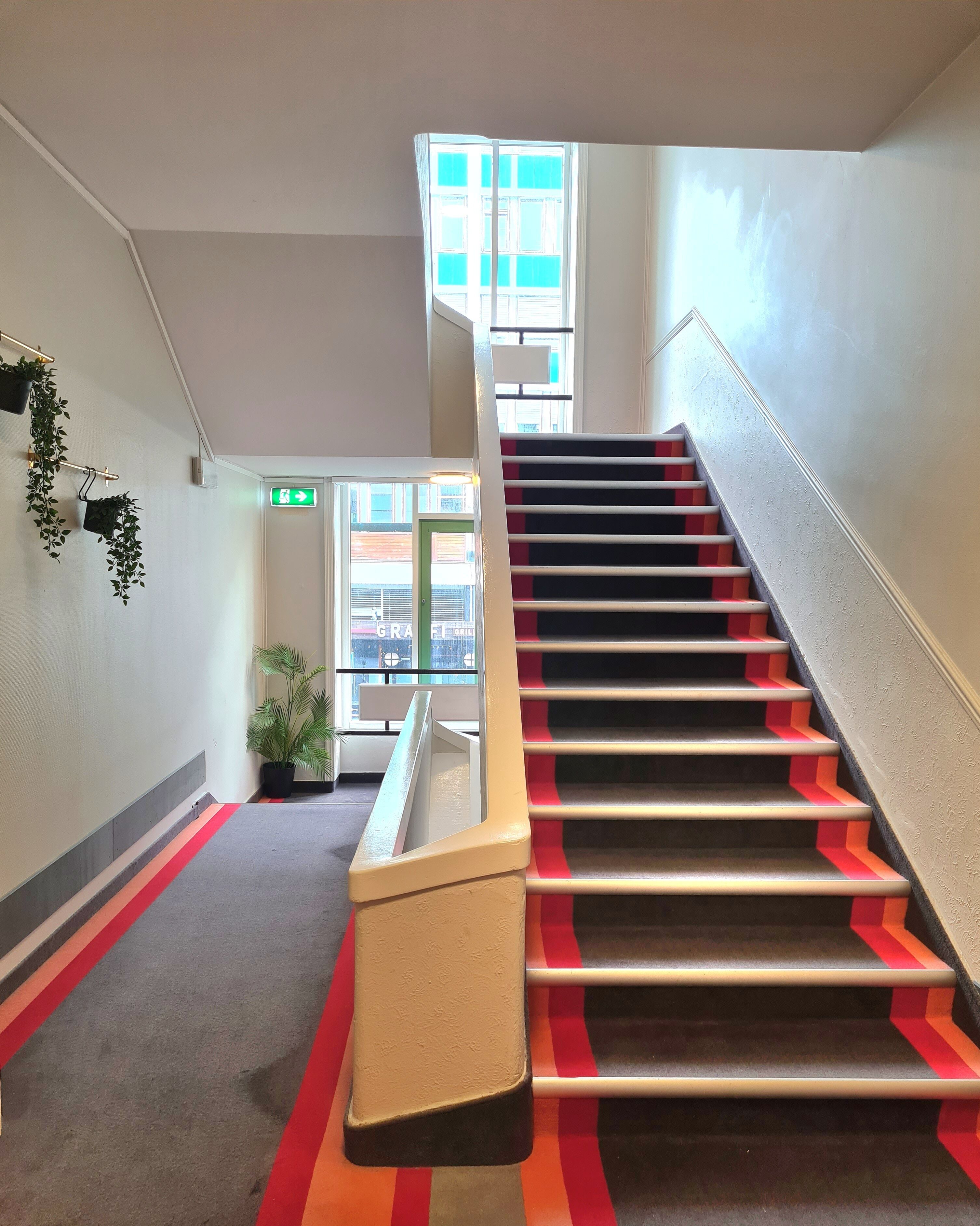 Una escalera con peldaños rojo y negro, una barandilla color beige y una planta a la izquierda.