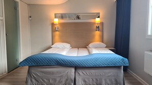 A double bed with a blue quilted bedspread and two pillows, flanked by two wall-mounted light fixtures, in a room with light wooden flooring and a view of a blue curtain. - City Living Sentrum Hotell (Trondheim)