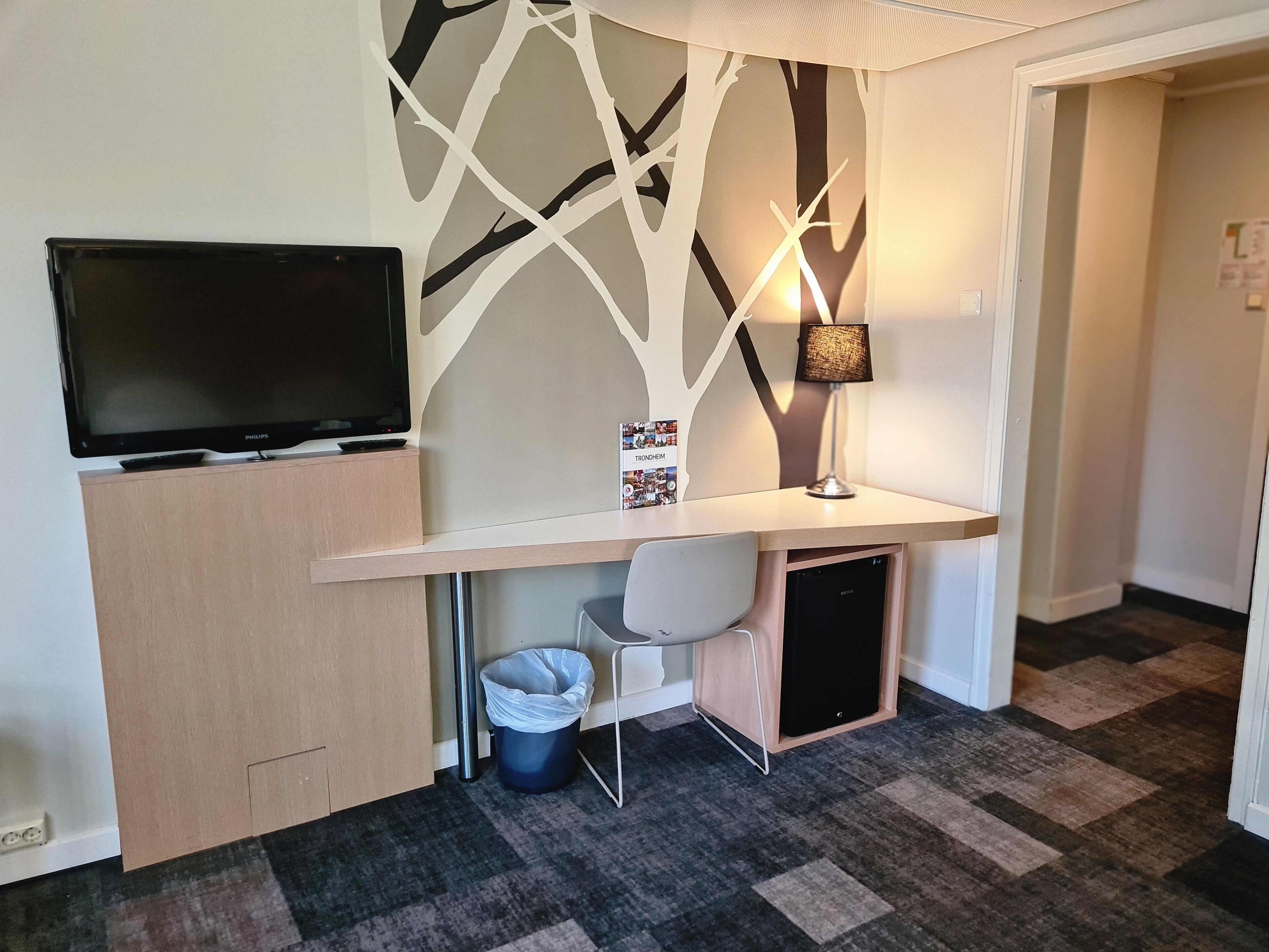 Ein Hotelzimmer mit Schreibtisch, Fernseher, Lampe, Stuhl und Mülleimer.