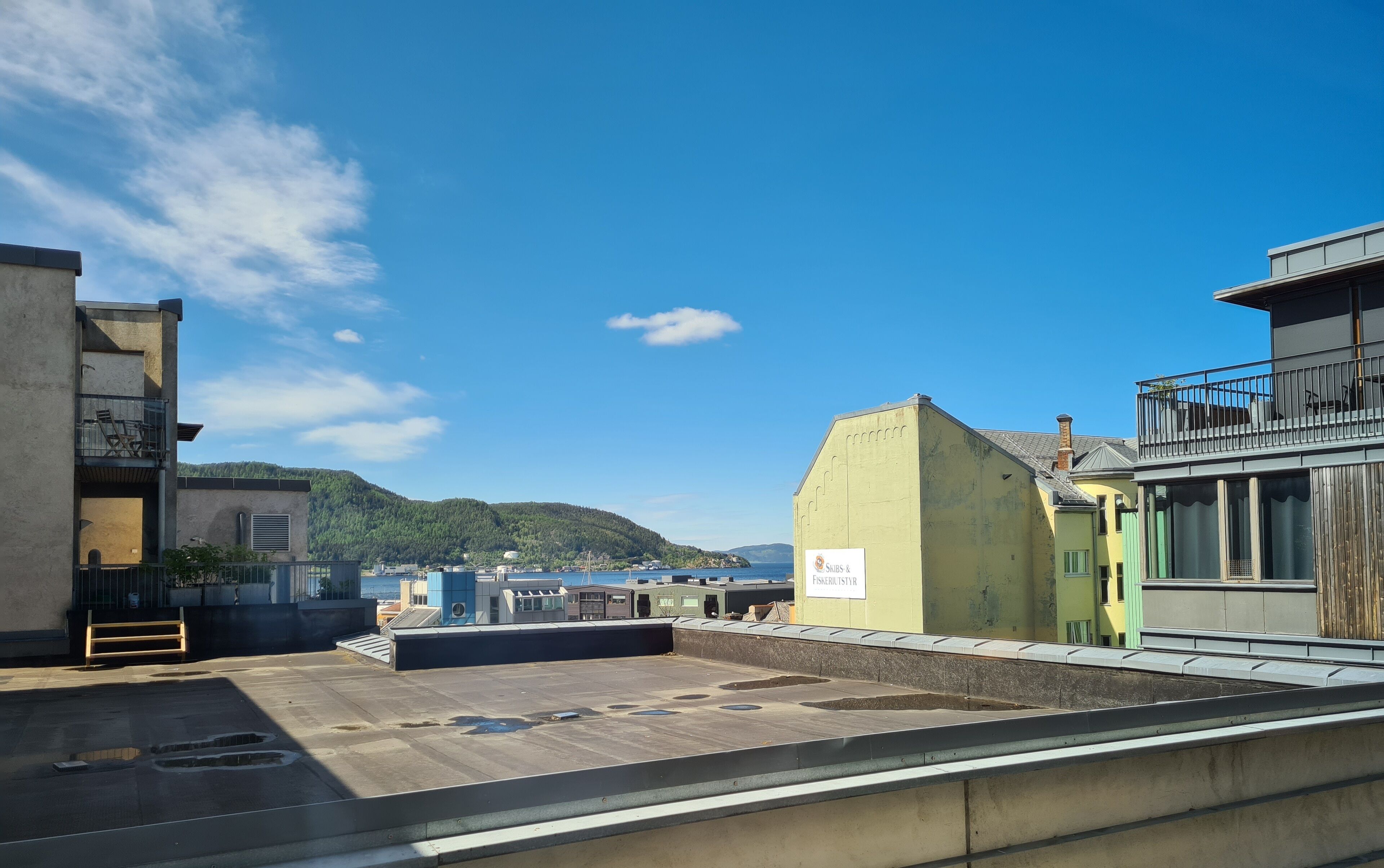 Eine Dachterrasse mit Blick auf Meer und Hügel, mit einem gelben Gebäude und einem Schild mit der Aufschrift 'Skibs & Fiskefabriksver'.