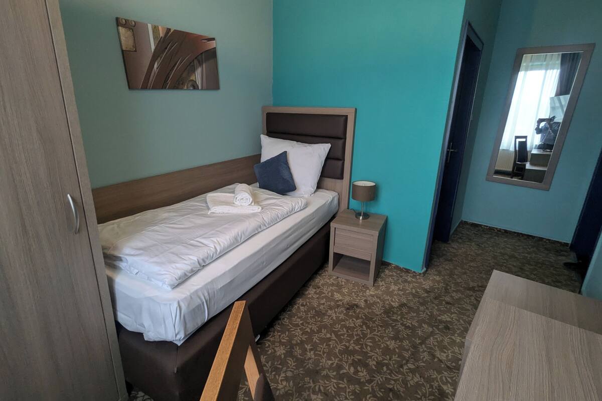 Un lit bien fait, avec du linge de lit blanc et une serviette pliée, une table de chevet avec une lampe, un miroir au mur et une porte sur la gauche.