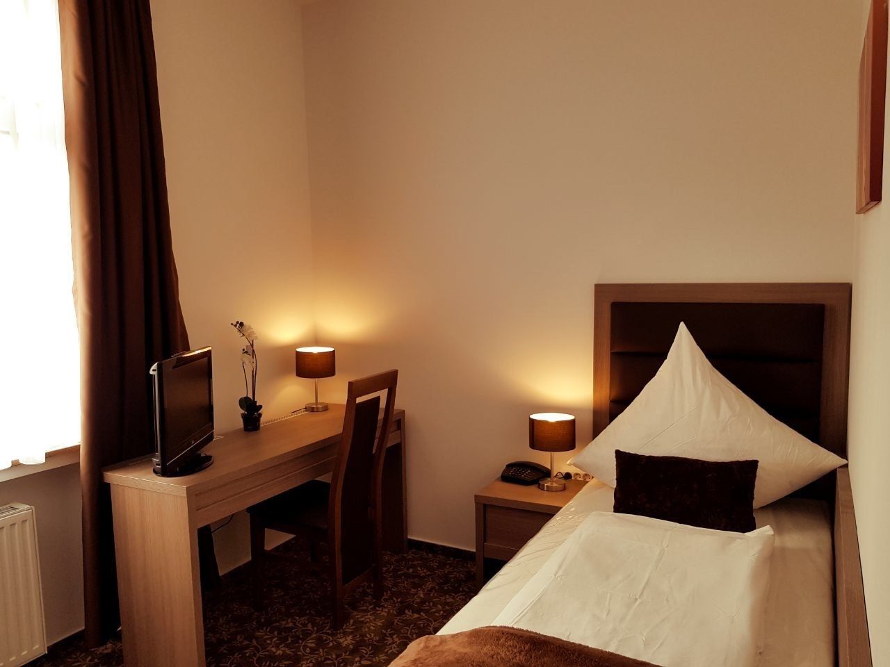 A hotel room with a bed, desk, chair, lamps, and a television.