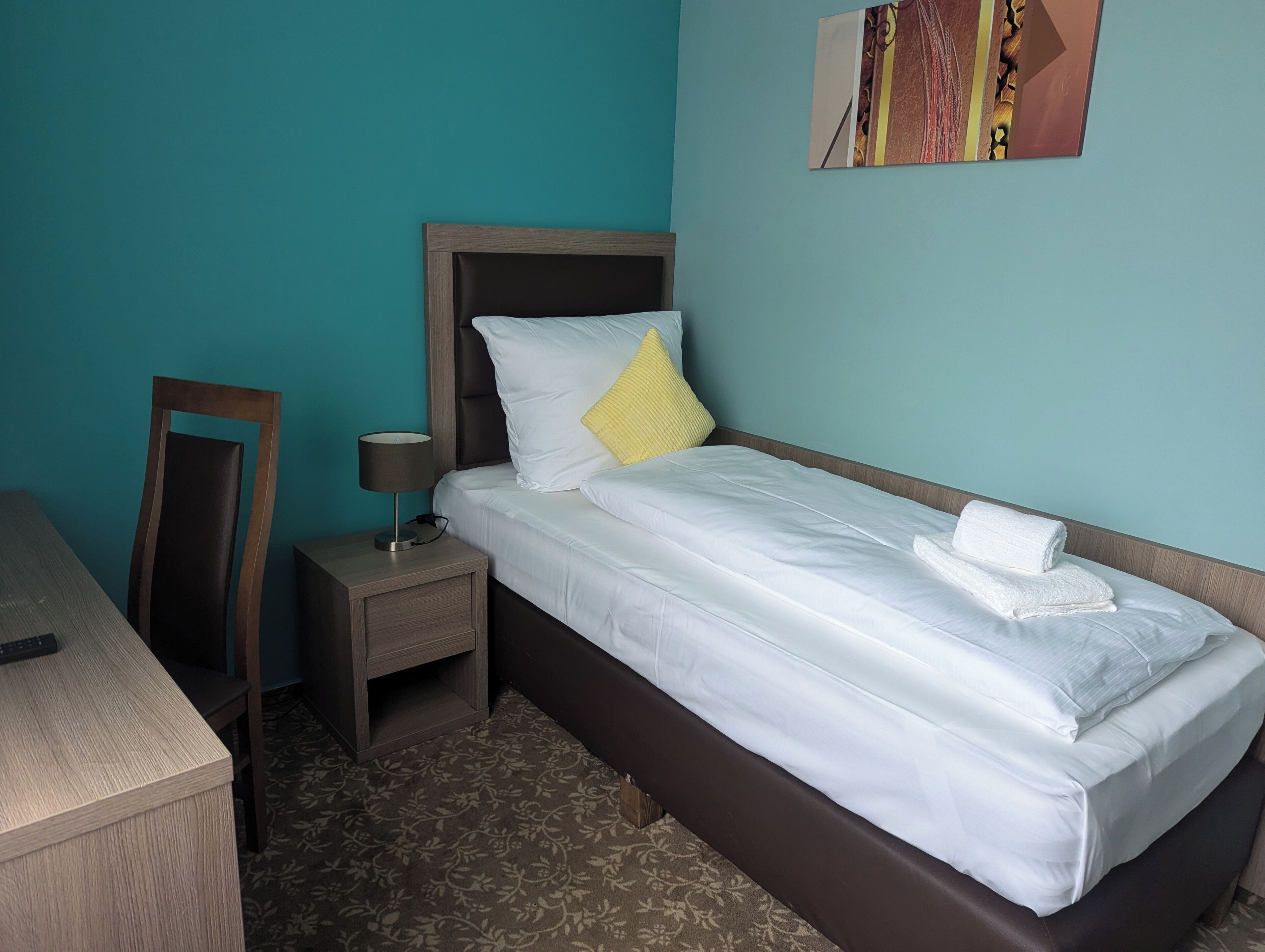 A neatly made bed with white linens and a yellow pillow, a bedside table with a lamp, and a chair against a teal wall.