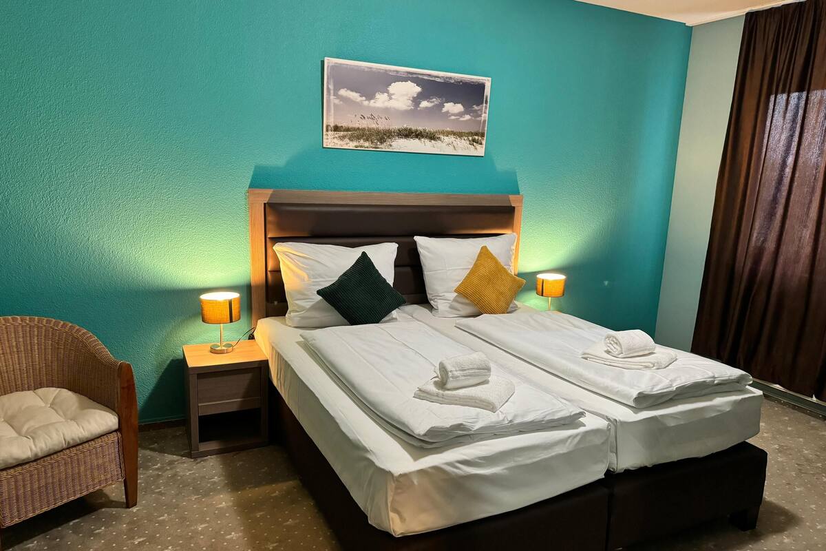 Une chambre d’hôtel avec un lit, deux oreillers, une table de chevet, une lampe, une chaise et un tableau au mur.