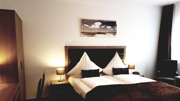 A hotel room with a bed, two bedside tables, a nightstand, a chair, a desk, and a wall-mounted picture. - easy&cosy Hotel Moers (Moers)
