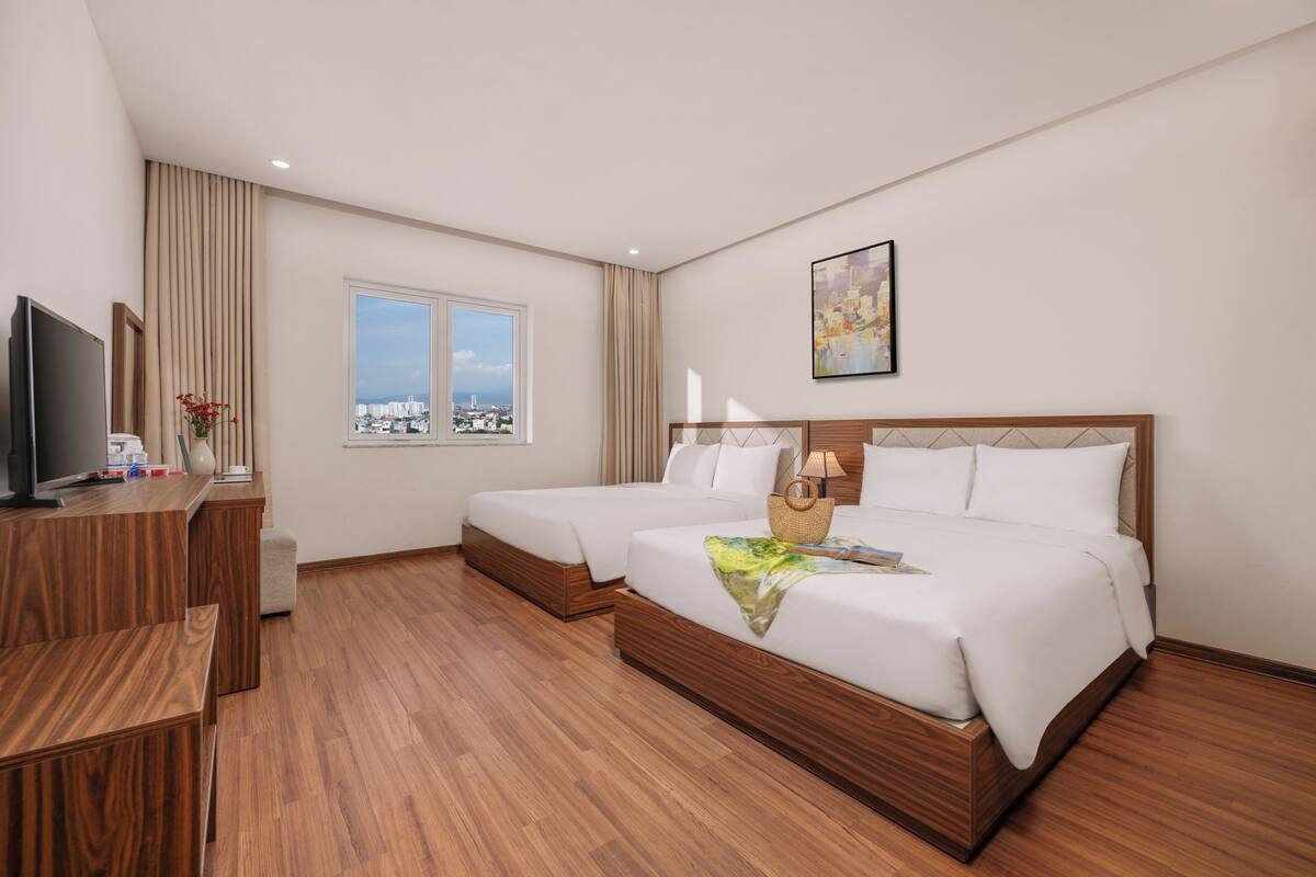 Habitación de hotel con dos camas, un escritorio, un televisor, una ventana con vista a la ciudad y un cuadro en la pared.