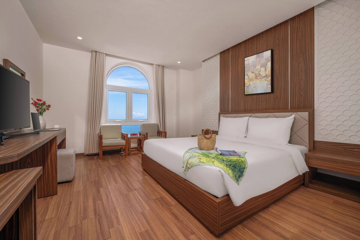 Habitación de hotel con una cama de madera, un escritorio con televisor, una silla y vista al mar a través de un ventanal grande.