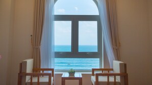 Una habitación con un amplio ventanal de arco que da al mar, dos sillas de madera, una mesita con una planta y cortinas color beige.