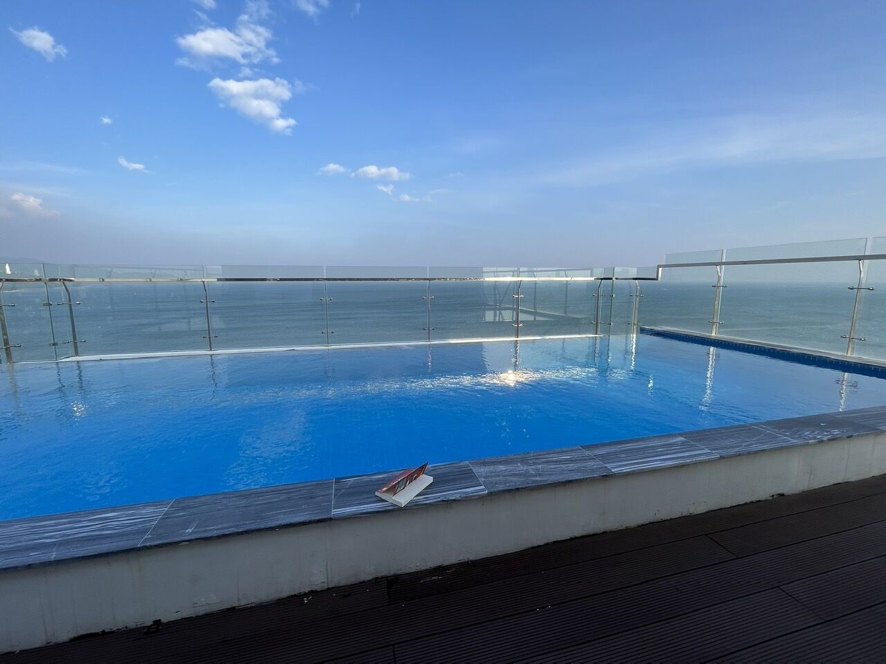 A clear blue pool with a glass barrier, surrounded by a wooden deck, overlooking a calm sea.