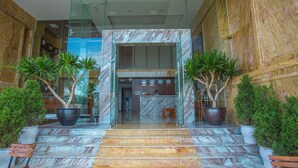 Una escalera de mármol conduce a una recepción con un recepcionista, plantas en macetas y un banco.