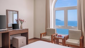 Habitación de hotel con un ventanal grande, un escritorio con una computadora portátil, un televisor, dos sillas, una mesita con fruta y una cama.