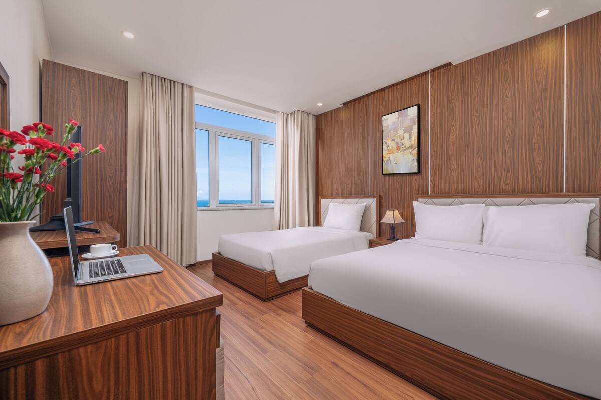 Habitación de hotel con una cama grande, una cama más pequeña, un escritorio con una computadora portátil, un florero con flores y una ventana con vistas.