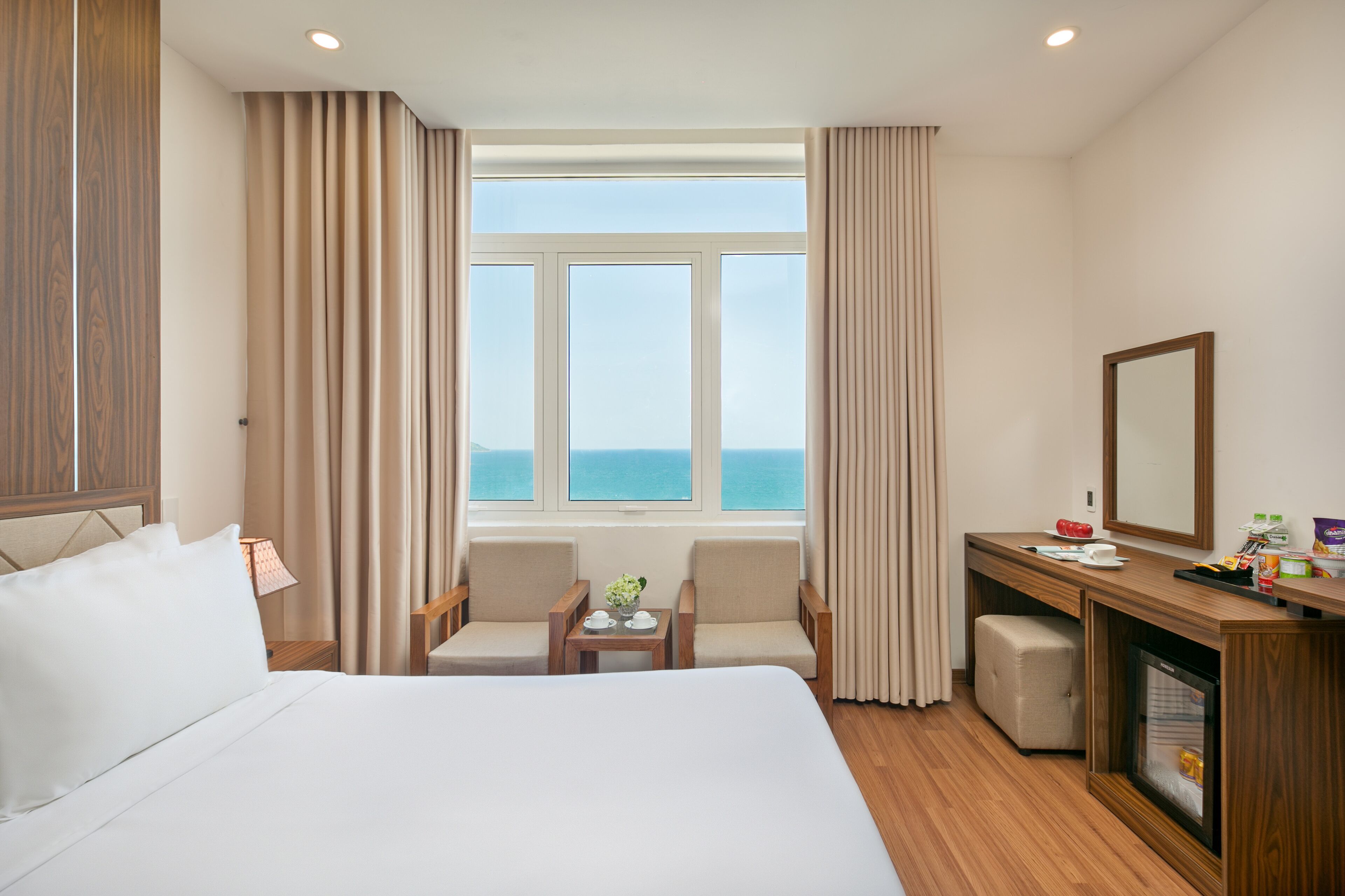 A hotel room with a large bed, two armchairs, a small table with a plant, a desk with a mirror, and a view of the sea through a large window.