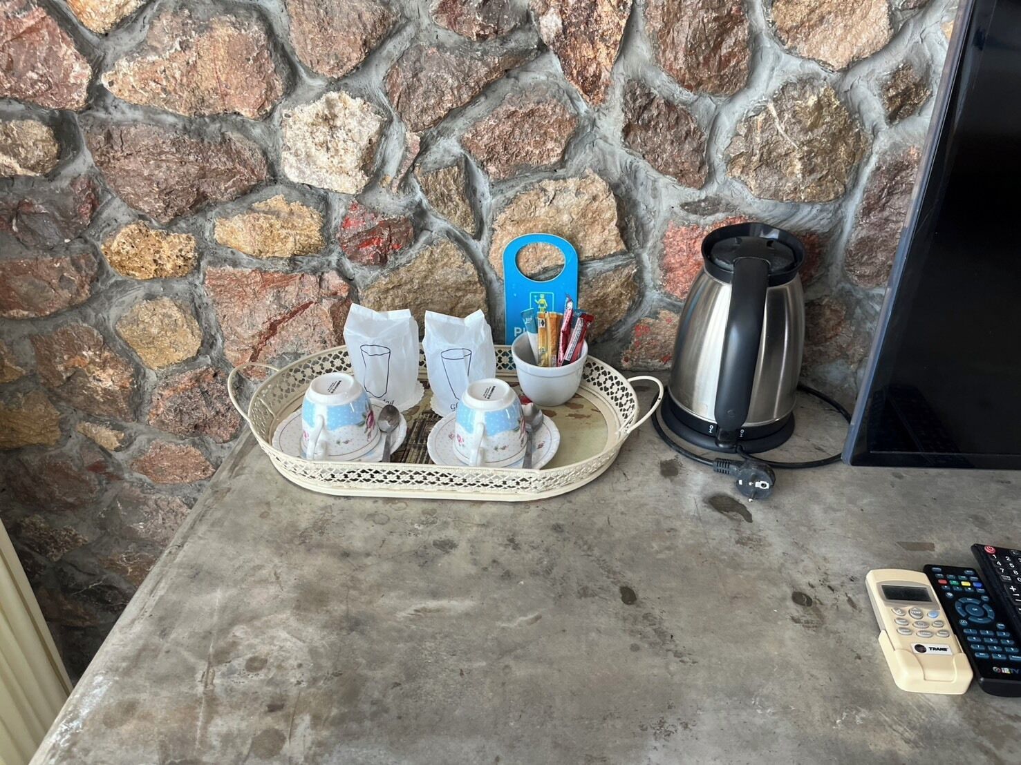A stone wall, a tray with teacups and sugar packets, a kettle, and a mobile phone on a table.