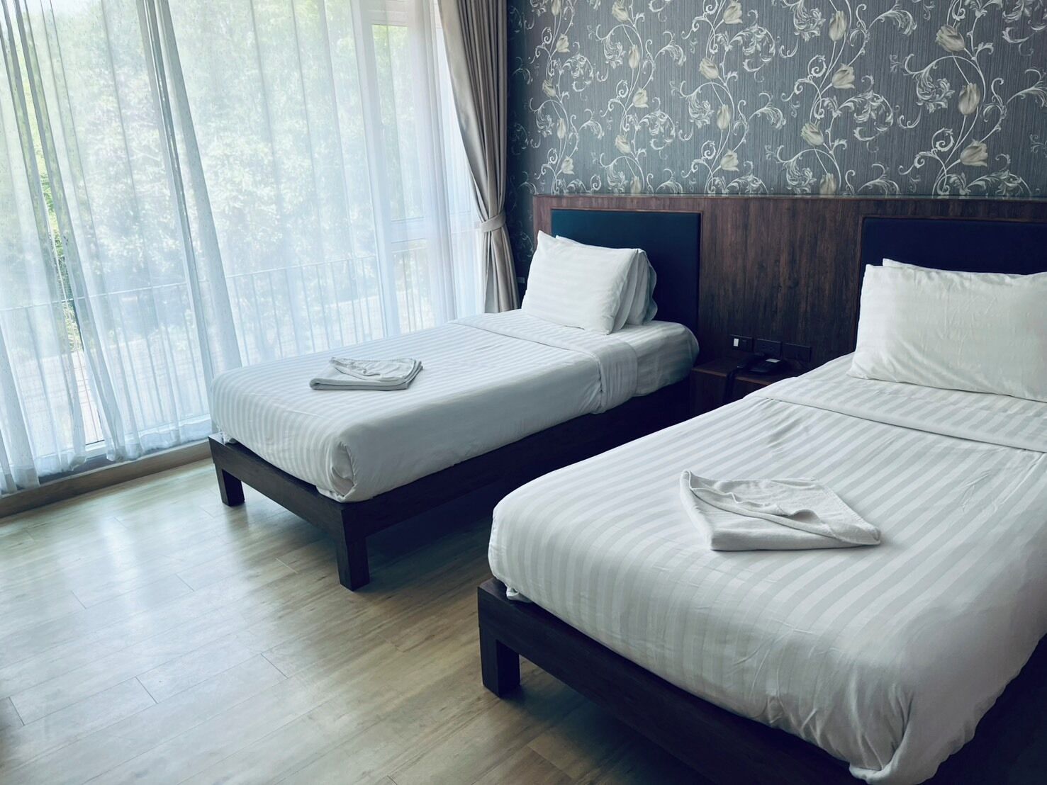 Une chambre d’hôtel avec deux lits, une tête de lit en bois, un mur à motifs et une grande fenêtre avec des rideaux légers.