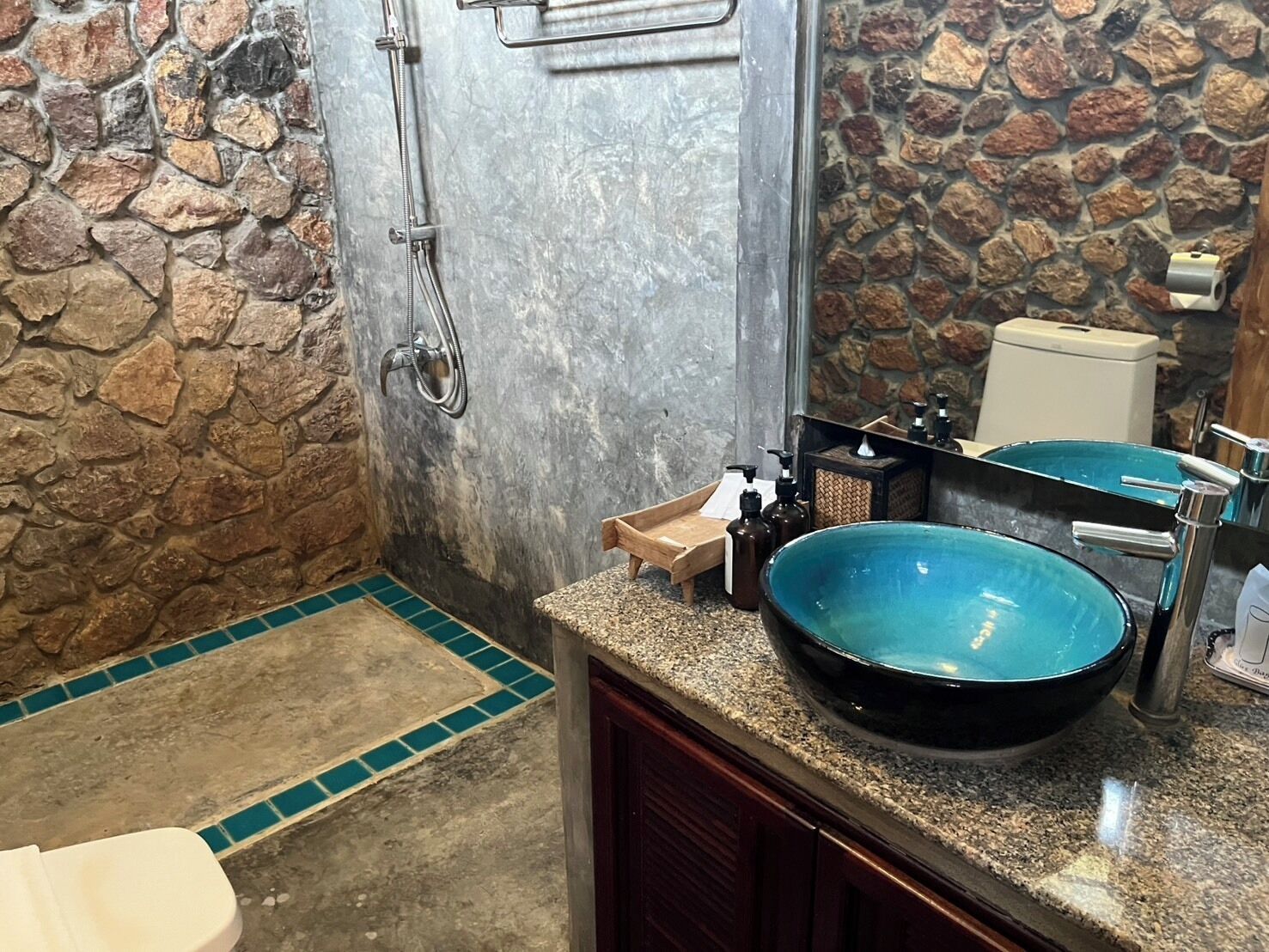 A bathroom with a stone wall, a shower, a sink with a blue basin, and a toilet.