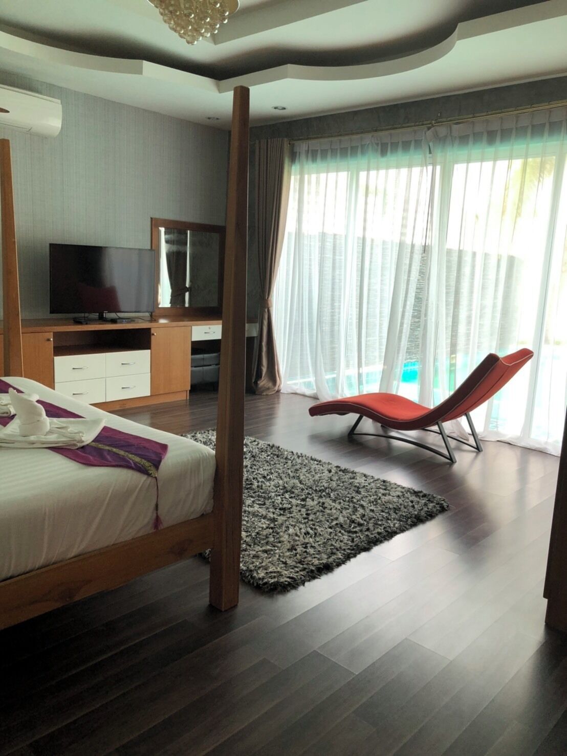 A modern bedroom with a four-poster bed, a flat-screen TV, a desk with a chair, and a large window with curtains.