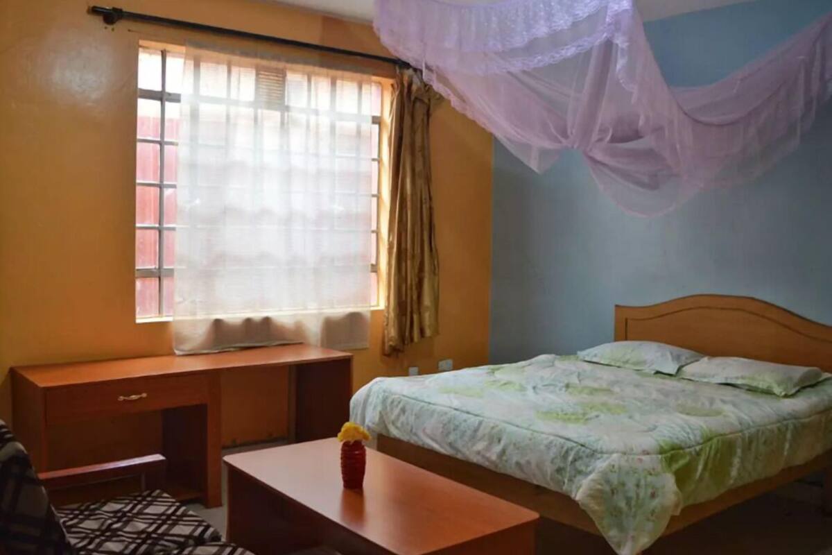 A bedroom with a bed, a desk, a chair, a window with curtains, and a mosquito net.